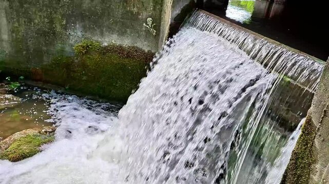 Fast flowing water cascades over a concrete sluice gate, creating foamy currents and dynamic textures. Rustic industrial structure meets nature in a peaceful countryside setting