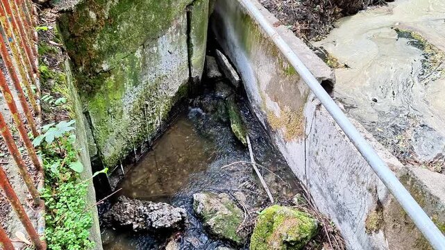 Water flows through a narrow concrete channel beside moss covered stone walls and natural rock formations. Gentle currents, textured surfaces, and a peaceful outdoor setting create a calm countryside 