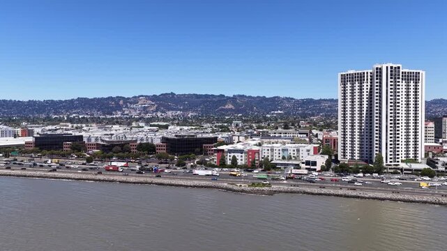Drone view flying over the San Francisco Bay looking at the Emeryville waterfront and skyline with heavy traffic on the Eastshore Freeway and the Berkeley Hills in the background on a sunny day