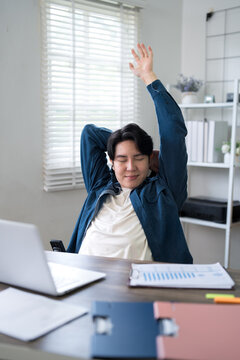 Young Asian professional stretching arms at desk with laptop taking wellness break from remote work at home office
