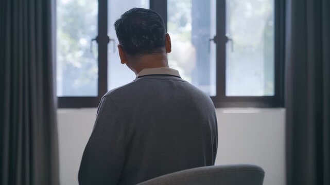 Back view of lonely elderly grandfather man sitting in wheelchair looking at the window closed with curtain for thinking or nostalgia. Retirement, dementia, oldness, disability, nursing home concept