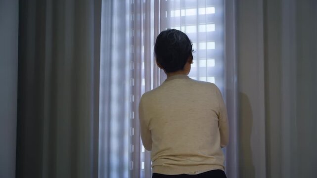 Back view of lonely grandmother woman stands by window closed with curtain thinking or nostalgia, gazing thoughtfully into the distance. Retirement, dementia, oldness, disability, nursing home concept
