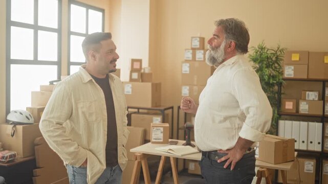 Two men with packages and arms crossed, smiling among stacked boxes and shelves inside a building shipping office; confidence teamwork.