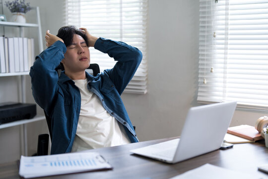 Young Asian businessman feeling stressed and overwhelmed while working on laptop at bright modern office desk