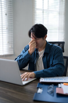 Young Asian man experiencing work stress rubbing eyes while working on laptop at home office desk