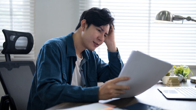 Stressed young Asian businessman holding document with worried expression while working at modern office desk