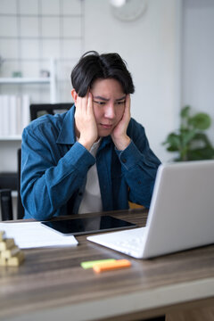 Stressed young Asian man holding head in frustration while working on laptop at home office desk