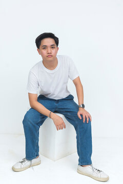 A young Southeast Asian man sits on a white cube in a studio setting, facing forward with a serious expression and relaxed posture.