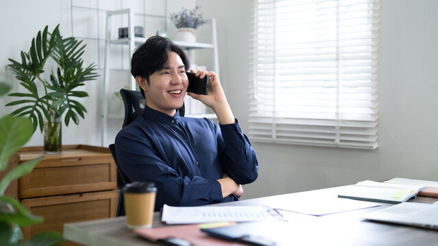 Young Asian businessman laughing on phone call while working from bright home office with coffee and documents