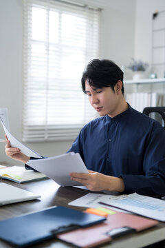 Young Asian professional reviewing business documents at desk in bright home office workspace
