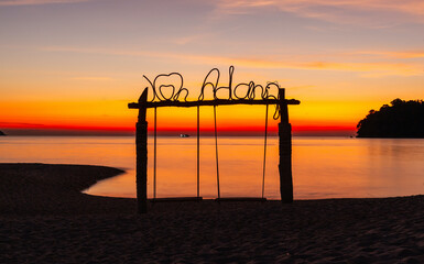 Fototapeta premium Sunset view with Koh Adang sign on beach. Calm ocean waters and vibrant sky. Silhouette of swings and tropical island landscape. Thailand coastal scenery at dusk.