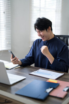 Young Asian businessman celebrating success with fist pump while reading smartphone message at modern office desk