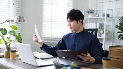 Young Asian professional reviewing paperwork and digital tablet while working from home office with...