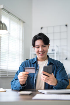 Young Asian man making smartphone payment with credit card while working remotely at bright home office desk