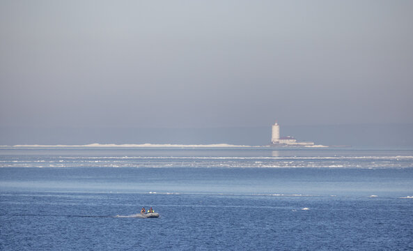 A small inflatable boat with fishermen crosses a calm, icy winter Baltic sea toward a distant Tolbukhin lighthouse on the horizon, creating a tranquil, minimalist coastal scene