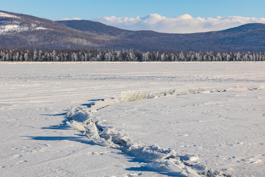 View of the frozen surface of Lake Baikal with ice cracks and snow-covered mountains in the background in Davsha, Buryatia, Russia.