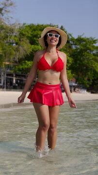 Smiling Asian woman wearing a red bikini with a matching skirt and a straw hat while walking forward in the shallow sea water