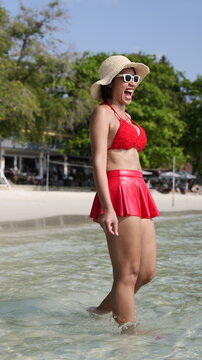 Cheerful Asian woman in a red bikini and skirt wearing a straw hat and sunglasses while wading in the clear ocean water at the beach