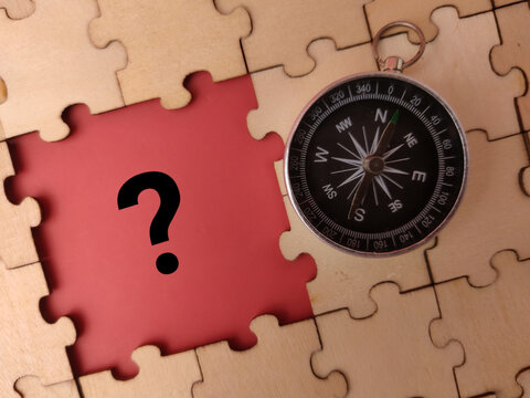 A circular magnetic compass rests on a wooden jigsaw puzzle next to a missing piece showing a question mark