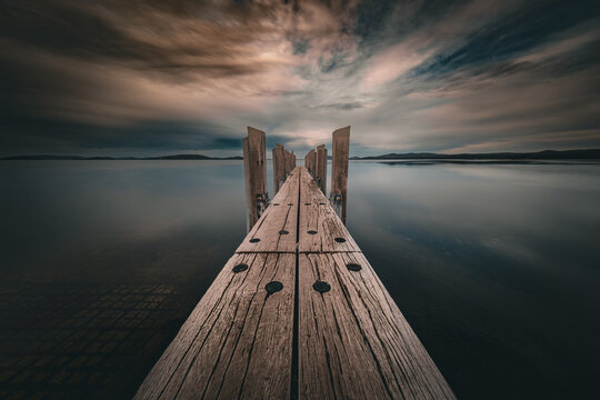 View of the wooden jetty at the Princess Royal Sailing Club extending into the calm waters of the bay under a dramatic, cloudy sky in Little Grove, Western Australia, Australia.