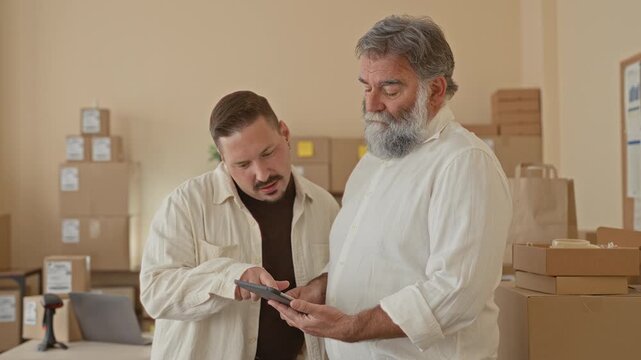 Two men pointing at a tablet with hands among stacked packages and scanner in a packing building; teamwork success.