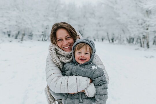 Smiling mother embracing her young son in a snowy winter park, featuring soft pastel tones and cozy knitwear for family bond and seasonal lifestyle content