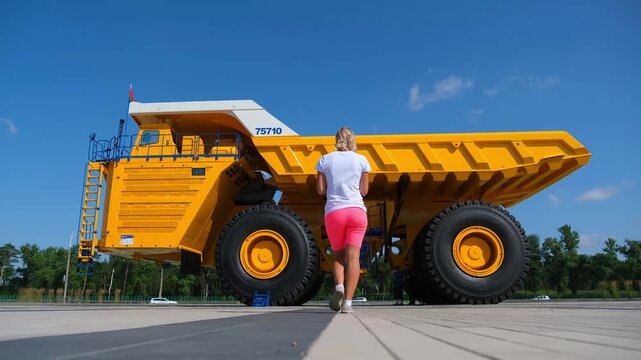 Minsk. Belarus. 09.08.2025 Woman standing beside giant yellow mining dump truck. Forced perspective photography creates illusion of tiny human near heavy machinery. Engineering scale contrast.