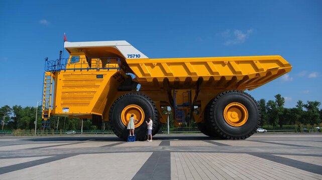 Minsk. Belarus. 09.08.2025 Woman and man standing next to giant yellow haul truck in outdoor area. Industrial machinery and mining vehicle exploration for transport tourism and display.