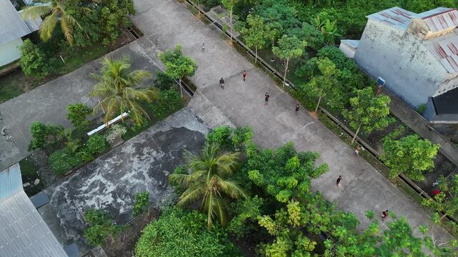 An aerial view captured by a drone shows poor rural children playing and throwing things at the camera on a concrete path surrounded by tropical trees in a village in Indonesia.