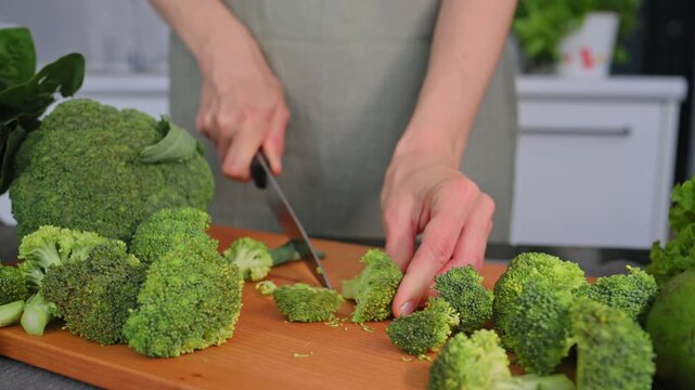 Woman cutting organic broccoli on wooden board for homemade meals. Fresh broccoli, green vegetable, farm produce communicates healthy nutrition, everyday cooking routine and commercial food