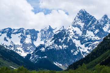 Obraz premium Snow-covered mountain peaks tower against a cloudy sky, with green valleys and rocky terrain creating a stunning natural landscape