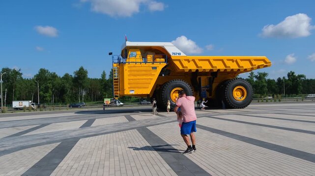 Man spins a child around standing next to a massive yellow mining truck. Father and daughter play outside near heavy machinery during a sunny summer day out.