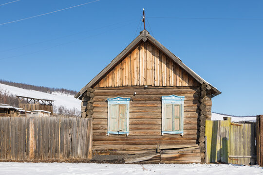 View of a traditional wooden log house with light blue window frames in a snowy rural landscape under a clear blue sky in Baikalskoye, Buryatia, Russia.