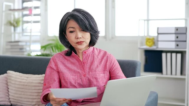 Older woman working on laptop reading document that made her frustrated, financial troubles, health concerns, bank debt notification, unexpected bills, feel distressed