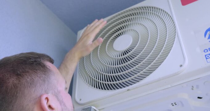 Close-up of a man inspecting and testing an indoor air conditioning unit by placing his hand near the fan grille to check airflow, cooling performance, and ventilation system operation.