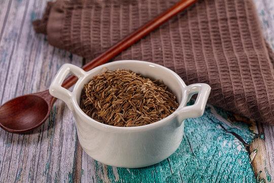 Dry cumin seeds in ceramic bowl with wooden spoon on textured wood background