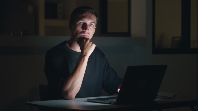 Programmer coding at night on laptop then stopping and looking at camera thinking about solution. Coding developer problem solving, software engineer focused in dark workspace with idea moment