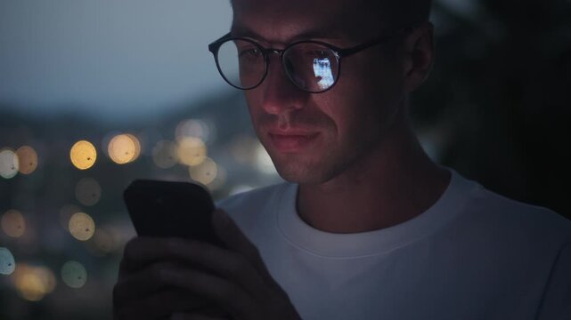 Smartphone user scrolling social media at night as screen reflects in glasses. Close up man portrait highlights mobile addiction online communication technology and digital lifestyle in urban lights.
