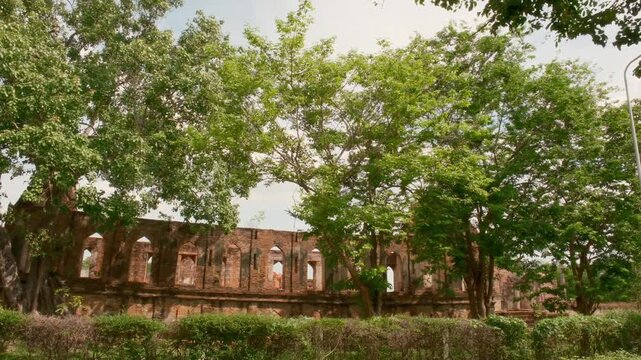 Wide exterior view of the ancient red brick ruins at Wat Kudi Dao in Ayutthaya, Thailand. Large green trees and a manicured hedge partially obscure the long historic assembly hall under a bright sky.