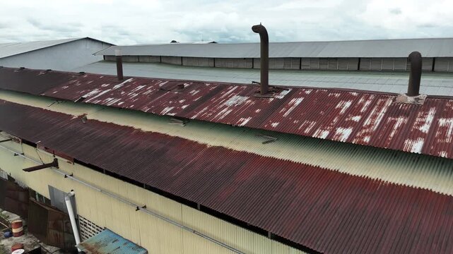 A drone view showing the old, rusty, corrugated factory roof with its smokestack, contrasting with the newer, cleaner gray metal warehouse roof.