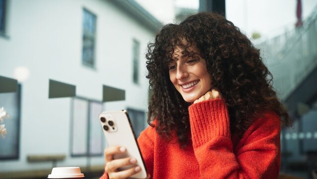 Caucasian woman with curly hair smiling while looking at smartphone in hand, sitting indoors near window. Female in red sweater drinking takeaway coffee and browsing phone at cafe table.