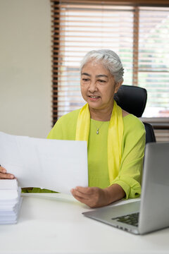 Old woman with grey hair work at home using notebook computer and reading document
