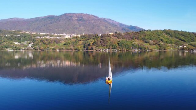 Aerial 4K drone orbiting a sailboat on scenic Lake Caldonazzo, surrounded by the majestic Trentino mountains with stunning reflections on the water surface.