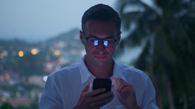 Mobile technology use at dusk as man scrolls smartphone feed with screen glowing in glasses. Innovation and connectivity concept showing modern tech lifestyle and constant online connection.