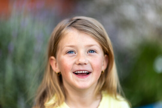 Young girl with long light‑brown hair, blue eyes, light yellow shirt, outdoors with blurred greenery