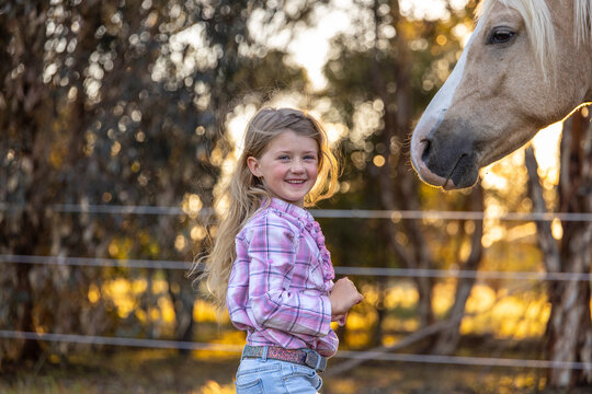 Caucasian girl standing near a light brown horse in fenced area with trees and golden sunlight