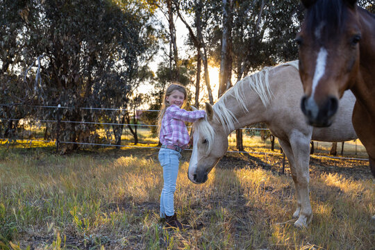 Young child in plaid shirt, jeans, boots, wide‑brimmed hat, gently petting a light‑coloured horse
