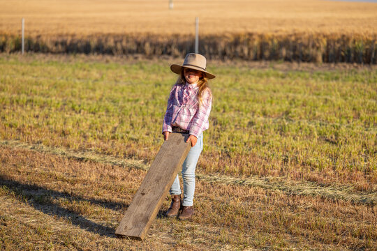 Young girl holding long wooden plank in a field with short grass and dry vegetation