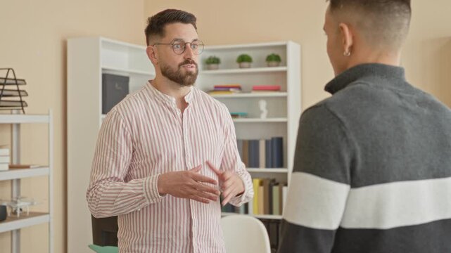 Man speaking with hand gestures and wearing glasses to a coworker in an office building, bookshelves visible and hands emphasized during discussion; teamwork focus.
