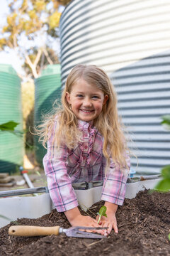 Young child with long blonde hair, leaning over raised garden bed and pressing soil around base